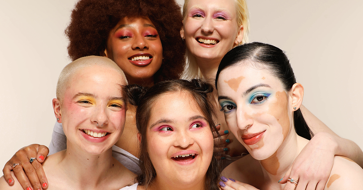 Image shows five stunning models smiling at the camera. Four of them present as female, and one presents as non-binary. They all have beautifully unique looks. Top right is a black model rocking an afro and mulberry pink glitter eye shadow. She has shimmering cheeks and deep red lipstick to match. Next to her is a model with albinism, she is wearing purple sparkly eyeshadow and has her shoulder length blonde hair slicked back. 

They have their arms around three models in front. The model on the far left has a blonde buzz cut and is beaming with yellow eyeshadow and bright pink blush. In the middle is an Asian model, she is showcasing some braces and is wearing hot cerise eyeshadow. She also has Downs Syndrome. The model on the far right has their hair slicked back in a long black ponytail. Their dark brown eyes are framed in shimmering cyan eyeshadow. This model has vitiligo which frames her features beautifully